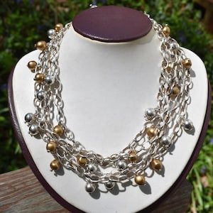 May include: A multi-strand necklace with silver chain links and alternating gold and silver-colored ball charms. The necklace is displayed on a white bust against a blurred green background.