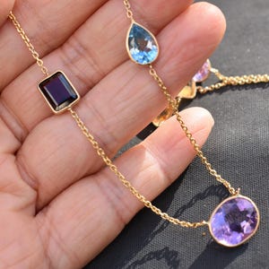 Peut inclure: Un collier en or délicat orné d'une pierre précieuse rectangulaire violet foncé, d'une pierre précieuse bleu clair en forme de goutte et d'une pierre précieuse violette ronde. Les pierres sont reliées par une fine chaîne en or.