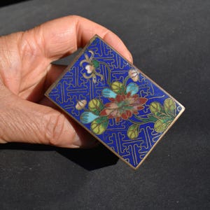 May include: A rectangular metal box featuring a vibrant blue enamel background with a gold-toned geometric pattern. The center displays a floral motif with red, green, and light blue enamel petals and leaves, framed by a thin gold-colored metal edge.