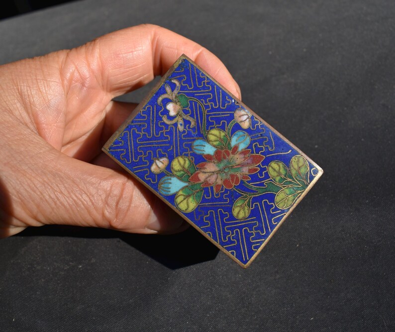 May include: A rectangular metal box featuring a vibrant blue enamel background with a gold-toned geometric pattern. The center displays a floral motif with red, green, and light blue enamel petals and leaves, framed by a thin gold-colored metal edge.