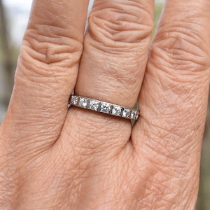 May include: A silver band with small, clear, square-shaped stones set in a continuous line around the band.