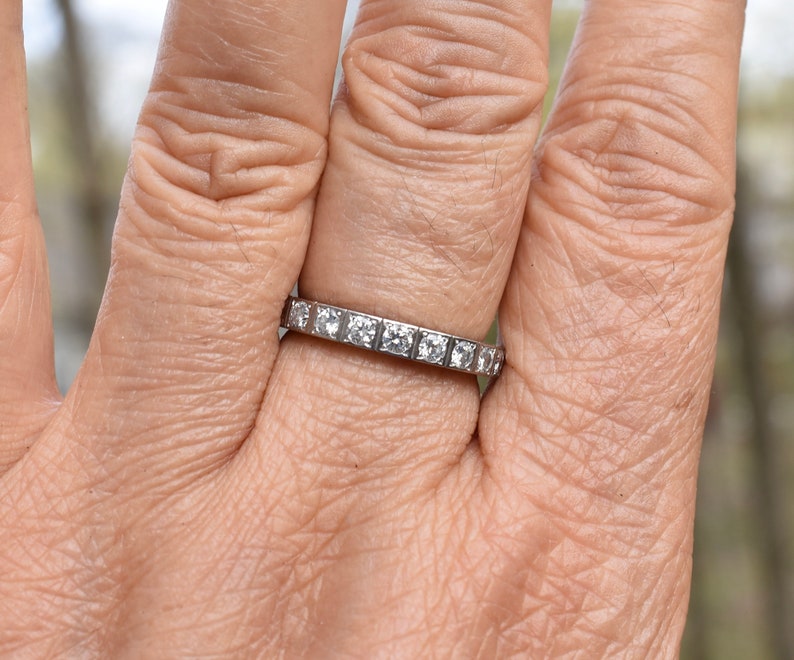 May include: A silver band with small, clear, square-shaped stones set in a continuous line around the band.