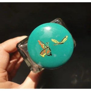 Könnte beinhalten: Tealfarbener Glasflaschenverschluss mit einem goldenen Schmetterlings- und Libellenmotiv.