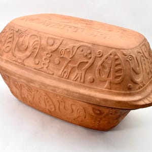 Könnte beinhalten: Ein ovaler Römertopf aus Terrakotta mit Deckel, verziert mit geprägten Reliefmustern von Hähnen, Bäumen und Schweinen. Auf der Oberseite des Deckels befindet sich der Schriftzug ROMERTOPF in erhabenen Buchstaben. Das Gefäß hat integrierte Griffe.