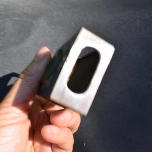 May include: A rectangular metal block with a smooth, brushed finish and a centered, elongated oval cutout. The object features a metallic silver tone with subtle hints of iridescent color variation along the edges and a hollow interior space.