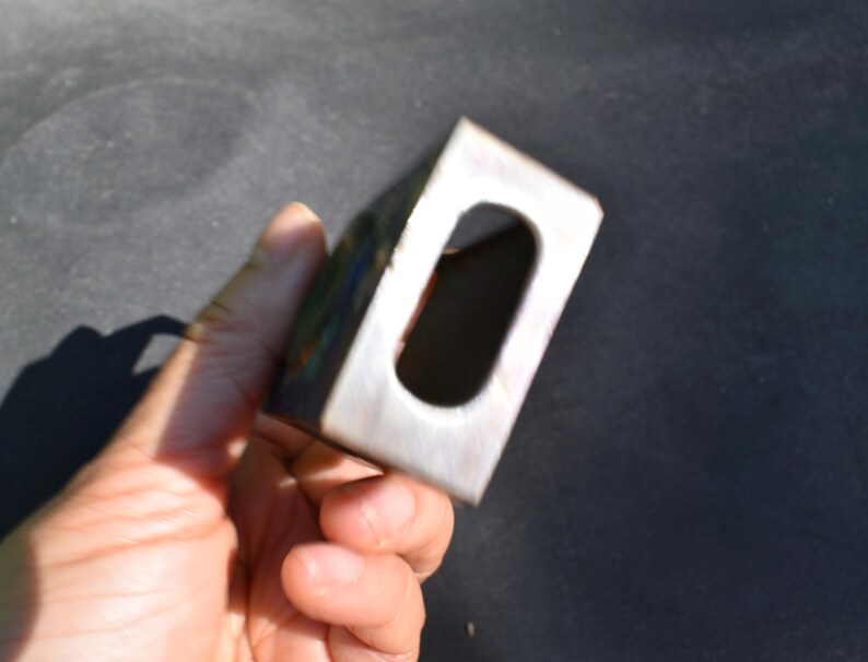 May include: A rectangular metal block with a smooth, brushed finish and a centered, elongated oval cutout. The object features a metallic silver tone with subtle hints of iridescent color variation along the edges and a hollow interior space.
