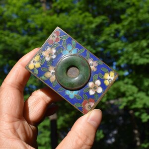May include: A rectangular metal plaque featuring a vibrant blue enamel background decorated with multi-colored floral patterns in red, yellow, and white. A circular, dark green stone ring with a central hole is mounted in the center of the piece.