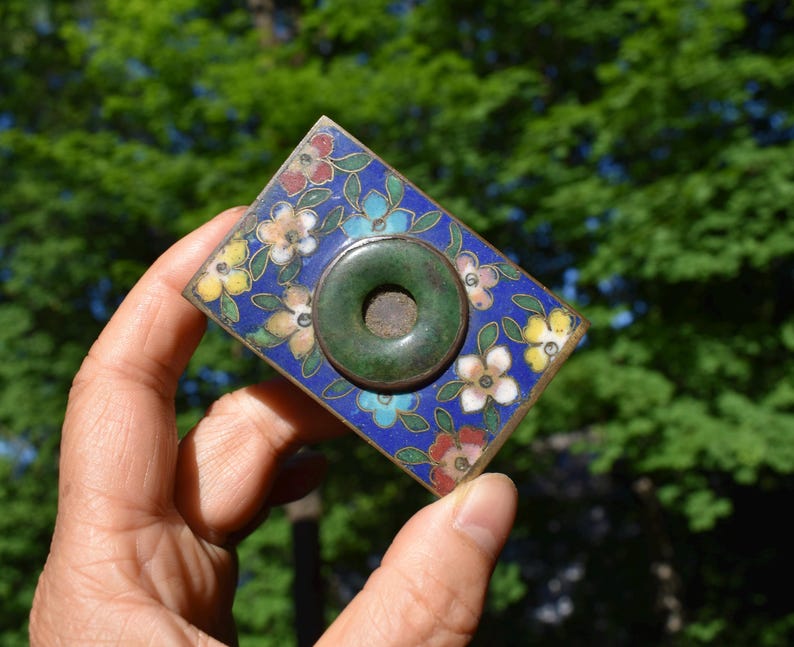 May include: A rectangular metal plaque featuring a vibrant blue enamel background decorated with multi-colored floral patterns in red, yellow, and white. A circular, dark green stone ring with a central hole is mounted in the center of the piece.