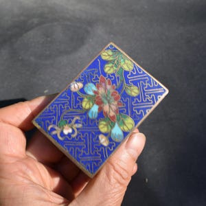 May include: A rectangular metal box featuring a vibrant blue enamel background with a gold-toned geometric pattern. The lid is decorated with a central floral motif in shades of red, green, and light blue, enclosed by a thin gold-toned metal border.