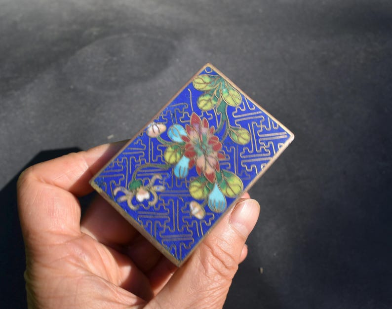 May include: A rectangular metal box featuring a vibrant blue enamel background with a gold-toned geometric pattern. The lid is decorated with a central floral motif in shades of red, green, and light blue, enclosed by a thin gold-toned metal border.