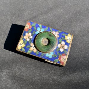May include: A rectangular metal box featuring a vibrant blue enamel surface decorated with multi-colored floral patterns in shades of pink, yellow, and light blue. A circular green stone disc is centered on the lid, framed by a thin metal border.