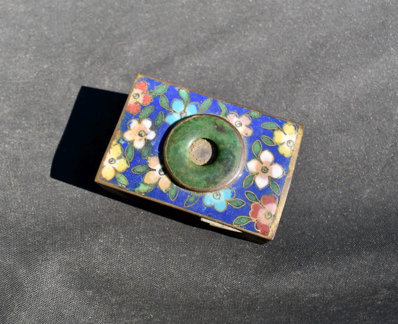 May include: A rectangular metal box featuring a vibrant blue enamel surface decorated with multi-colored floral patterns in shades of pink, yellow, and light blue. A circular green stone disc is centered on the lid, framed by a thin metal border.