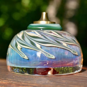 Puede incluir: Un frasco de perfume decorativo de vidrio con una tapa de latón. El frasco tiene una forma redondeada con un patrón arremolinado de colores blanco, verde y azul. La base del frasco es transparente y la parte superior es verde.