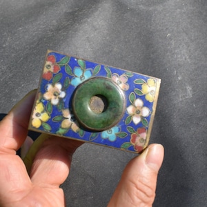 May include: A rectangular metal box featuring a vibrant blue enamel surface decorated with multi-colored floral patterns in red, yellow, and white. A circular, dark green stone ring is mounted in the center of the lid, creating a contrast.