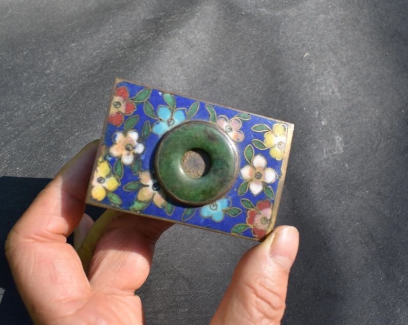 May include: A rectangular metal box featuring a vibrant blue enamel surface decorated with multi-colored floral patterns in red, yellow, and white. A circular, dark green stone ring is mounted in the center of the lid, creating a contrast.