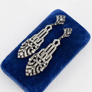 以下が含まれることがあります： 銀色の仕上げと透明なラインストーンが特徴の、アールデコ調のドロップイヤリングのペア。 イヤリングは、精巧なディテールを備えた細長い幾何学模様のデザインで、青いベルベットの表面に置かれています。