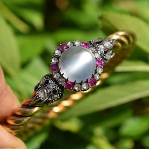 May include: A gold-toned bracelet with a twisted rope design. The bracelet features a central, round, light-colored stone surrounded by small, pink and clear stones. The ends of the bracelet have decorative, dark-colored accents.