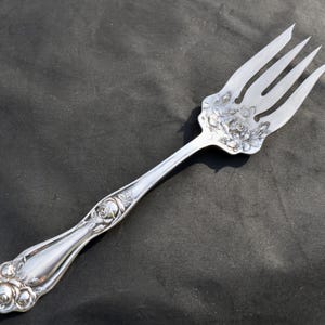 May include: Silver serving fork with a floral design. The fork has a detailed rose pattern on the handle and the tines' base. The tines are long and slender. The fork is made of a shiny, reflective metal.