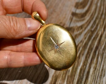 May include: A gold-colored oval locket with a starburst design and a small, clear gemstone at its center. The locket has a textured surface and a loop at the top for a chain. The locket is held in a hand.