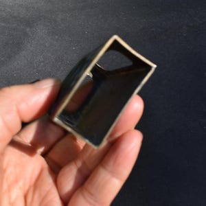 May include: A small, rectangular, hollow metal object with a dark, metallic finish. The item features an open-ended, box-like structure with a curved side and a circular cutout on one of the interior panels, showcasing a simple, industrial design.