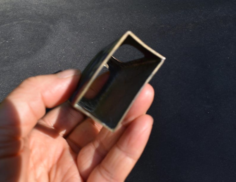 May include: A small, rectangular, hollow metal object with a dark, metallic finish. The item features an open-ended, box-like structure with a curved side and a circular cutout on one of the interior panels, showcasing a simple, industrial design.