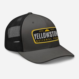 May include: A charcoal gray trucker hat with a black mesh back. The front of the hat features a yellow and white embroidered patch that reads "ESTD 1872 YELLOWSTONE NATIONAL PARK".