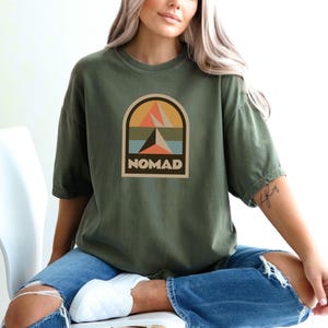 May include: Olive green t-shirt with a graphic featuring a mountain and arch design in orange, peach, blue, and brown. The word "NOMAD" is printed in a rectangular box below the design. The shirt has short sleeves and a relaxed fit.