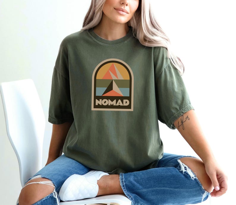 May include: Olive green t-shirt with a graphic featuring a mountain and arch design in orange, peach, blue, and brown. The word "NOMAD" is printed in a rectangular box below the design. The shirt has short sleeves and a relaxed fit.