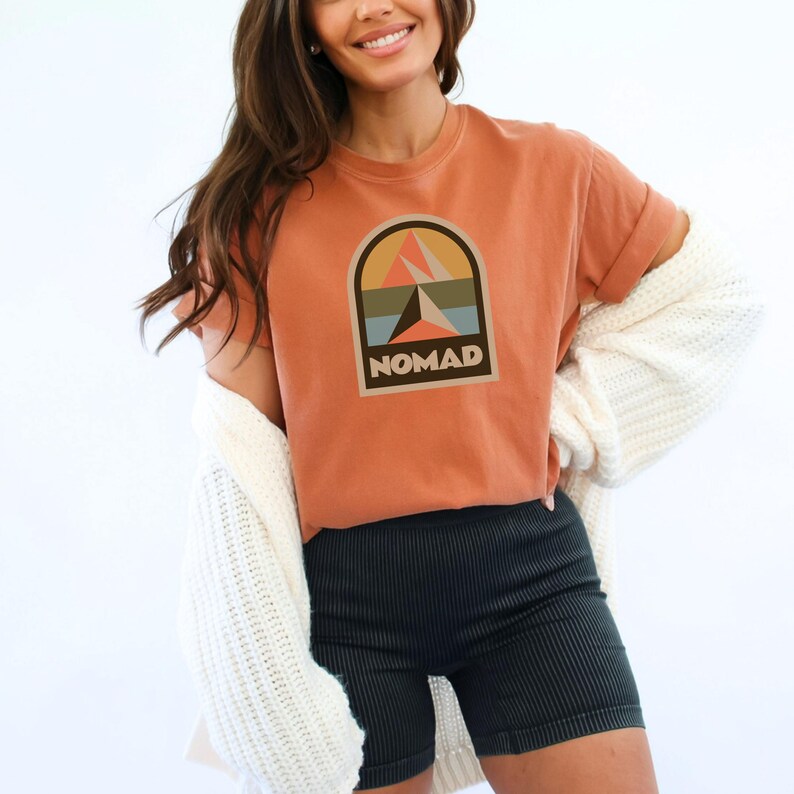 May include: Terracotta-colored t-shirt with rolled-up sleeves featuring a graphic design of a mountain scene within an arch, with the word "NOMAD" below. The shirt is paired with dark green ribbed shorts and a white cardigan.