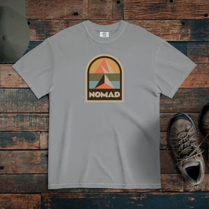 May include: A light gray t-shirt with a retro-style graphic that says "NOMAD." The graphic features a mountain scene in orange, teal, and beige. A green cap and hiking boots are also in the image.