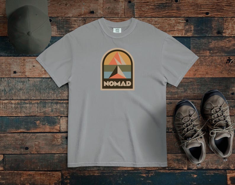 May include: A light gray t-shirt with a retro-style graphic that says "NOMAD." The graphic features a mountain scene in orange, teal, and beige. A green cap and hiking boots are also in the image.