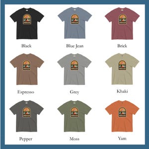 May include: Nine different colored t-shirts are displayed, each featuring a graphic with a mountain and sun design inside an arch, with the word "NOMAD" below. Colors include black, blue jean, brick, espresso, grey, khaki, pepper, moss, and yam.