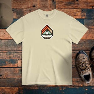 May include: A beige t-shirt with a colorful Basecamp logo featuring a tent design. The shirt is paired with a matching cap and hiking boots, suggesting an outdoor theme. The logo includes the word "BASECAMP".