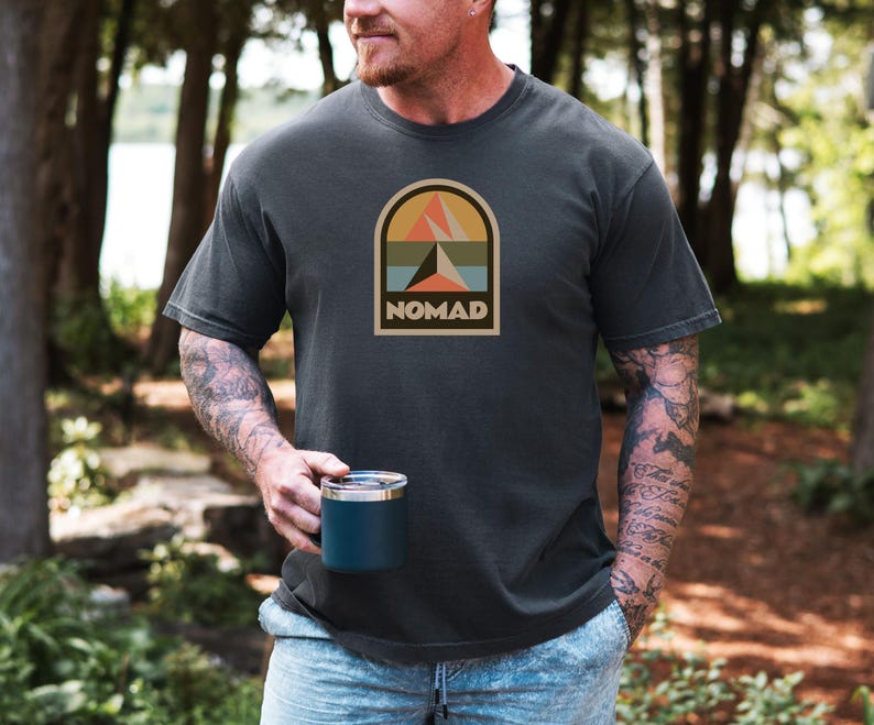 May include: Dark gray t-shirt with a retro-style graphic featuring a mountain scene and the word "NOMAD" in a rectangular arch. The person is holding a dark blue mug. The shirt is a comfortable, casual style.