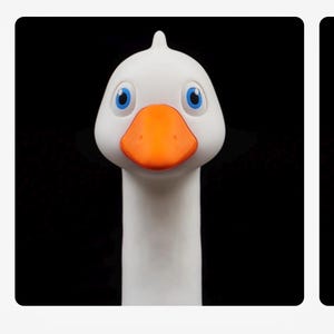 May include: A white goose head figurine with a bright orange beak and large blue eyes. The smooth, rounded head features a small crest at the top and a long, slender neck extending downwards, set against a solid, plain black background.
