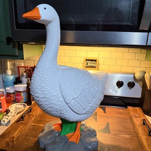 May include: A white goose figurine with a bright orange beak and feet, standing on a base of gray sculpted rocks and green grass. The goose has detailed molded feather textures on its body and a smooth, glossy finish on its head and neck.