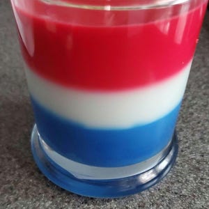 Pode incluir: Um frasco de vidro para velas com três camadas de cera vermelha, branca e azul. A vela está em um frasco de vidro transparente com uma base azul.
