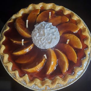 May include: A brown and white apple pie candle with a white whipped cream topping. The candle has a flaky crust and a glossy caramel sauce.