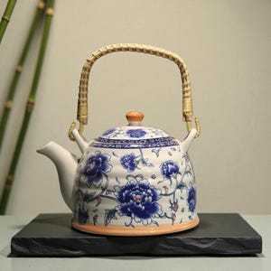 Könnte beinhalten: Weiße Keramik-Teekanne mit blauem Blumenmuster und einem geflochtenen Griff. Die Teekanne hat einen gebogenen Ausguss und einen Deckel mit einem orangefarbenen Knopf. Der Sockel hat eine orangefarbene Umrandung. Die Teekanne steht auf einer schwarzen quadratischen Oberfläche.