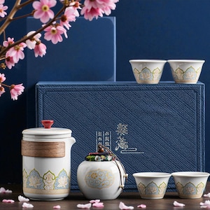 May include: A white ceramic tea set featuring a teapot, a small jar, and four teacups, decorated with a gold and blue floral design. The set is displayed on a blue box, with pink flowers in the background.