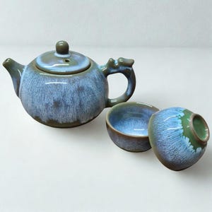 May include: A blue and green glazed ceramic tea set. The set comprises a teapot with a lid and two teacups. The teapot has a curved handle and spout. The teacups are small and round. The tea set is designed for serving tea.