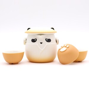 May include: A ceramic tea set with a panda design. The set includes a panda-shaped teapot with a lid, two small teacups, and a lid with a paw print. The teapot is white with black eyes and a black nose. The teacups and lid are orange.