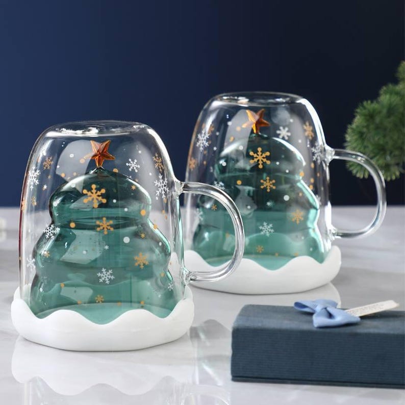 May include: Two clear glass mugs with a Christmas tree design inside. The tree is teal with a gold star on top. The mugs have a white base and are decorated with snowflakes and gold stars. A blue gift box with a blue bow is in the foreground.