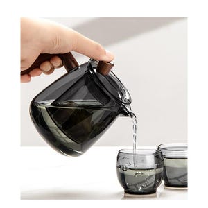Puede incluir: Una tetera de vidrio transparente con un asa de madera, vertiendo té en dos tazas de vidrio transparente. La tetera tiene un filtro integrado.
