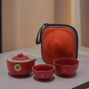 Peut inclure: Un service à thé en céramique rouge comprenant une théière, deux tasses et un étui de transport. La théière a un couvercle et un motif de pièce d'or. Les tasses ont un symbole doré. L'étui de transport est orange avec une poignée noire.