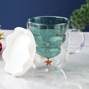 May include: A clear glass mug with a teal Christmas tree design, adorned with gold snowflakes and a star. The mug has a handle and a white, wavy-edged lid. The mug is designed for hot beverages.