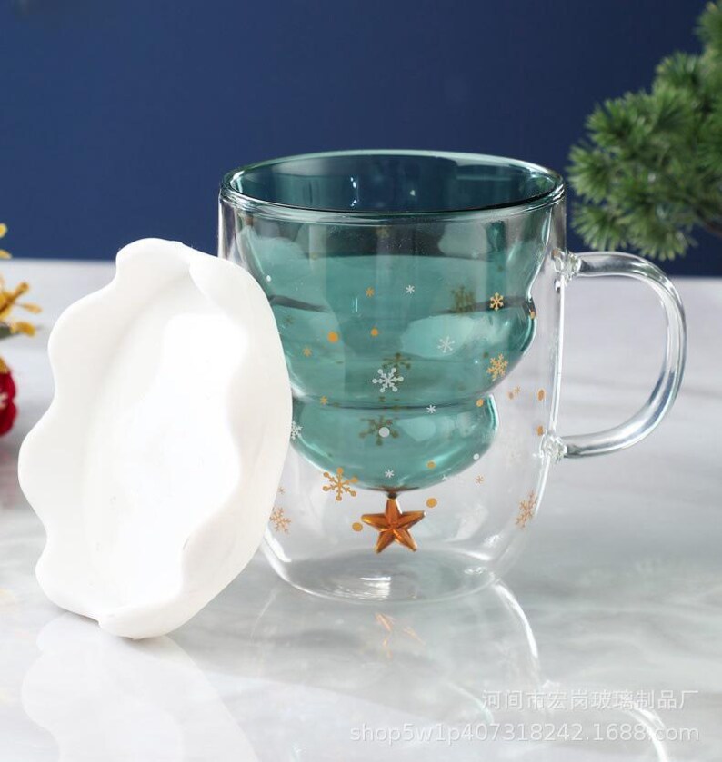 May include: A clear glass mug with a teal Christmas tree design, adorned with gold snowflakes and a star. The mug has a handle and a white, wavy-edged lid. The mug is designed for hot beverages.