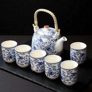 Könnte beinhalten: Ein weiß-blaues Teeservice mit floralem Muster. Das Set enthält eine Teekanne mit geflochtenem Griff und sechs passende Teetassen. Die Teekanne hat einen braunen Deckel und Ausguss. Die Tassen und die Teekanne sind auf einer schwarzen Oberfläche angeordnet.