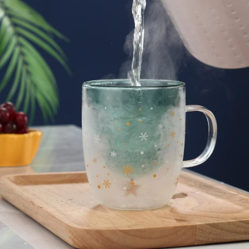 May include: A clear glass mug with a green ombre design and snowflake pattern is being filled with hot water. The mug sits on a wooden tray, with a small yellow bowl of red fruit in the background. Steam rises from the mug.