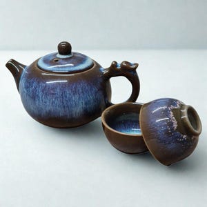 Könnte beinhalten: Ein Keramik-Teeservice mit brauner und blauer Glasur. Das Set enthält eine Teekanne mit einem griff in Drachenform und zwei passende Teetassen. Die Teekanne hat einen Deckel mit einem kleinen Knopf. Das Set ist zum Servieren von Tee bestimmt.